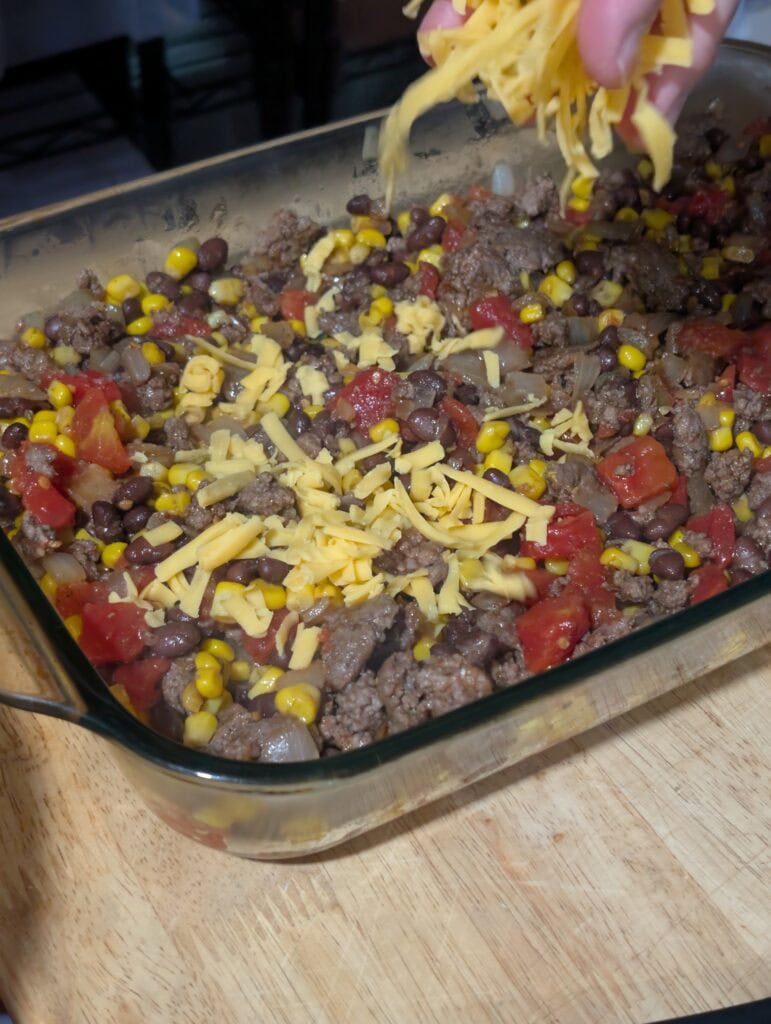 Hand dropping grated cheese over meat and tomatoes in a baking dish.