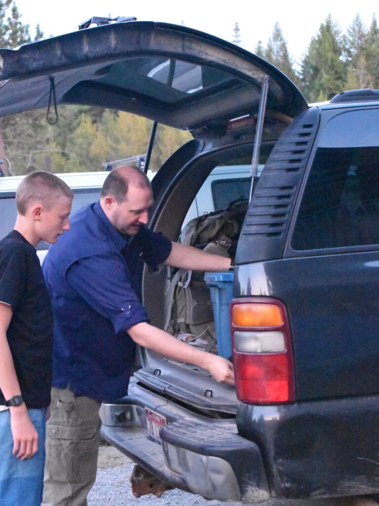 Dad and teenage son loading 72-hour kits and emergency supplies into the rear hatch of an SUV for a family evacuation drill.