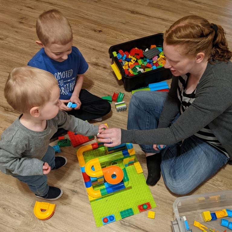 A mother and two young sons playing with colorful building blocks on a wooden floor, representing the quiet home life protected by a prepared mind.