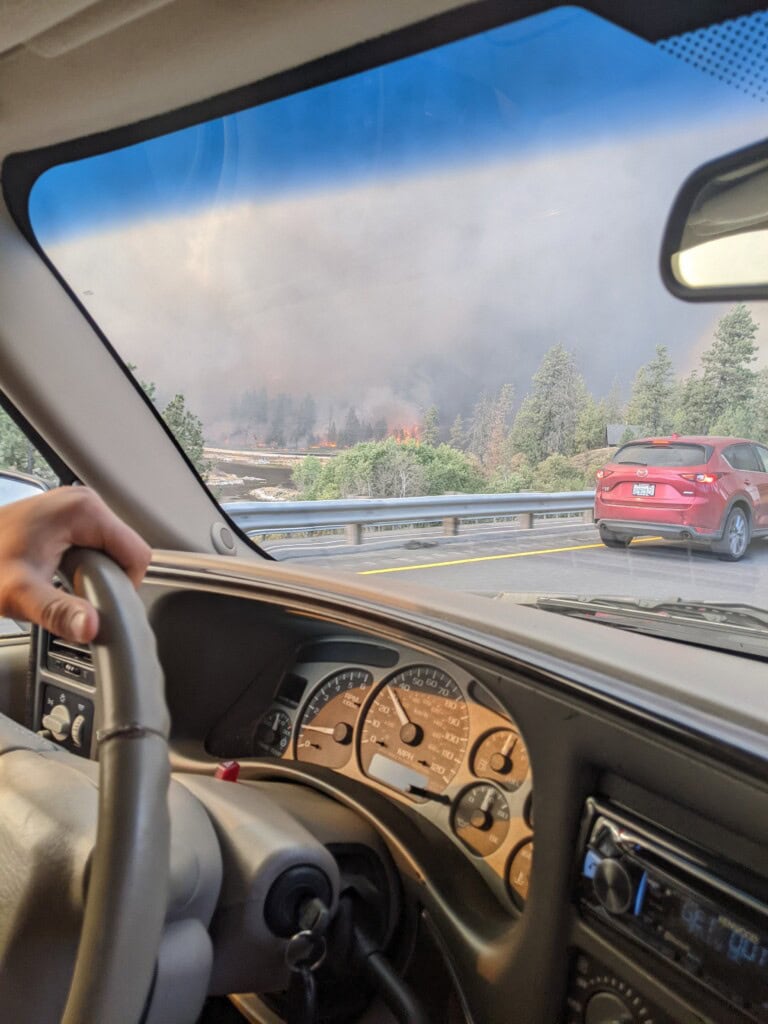 View from inside a car during a wildfire evacuation showing a driver's hand on the steering wheel and smoke on the horizon.
