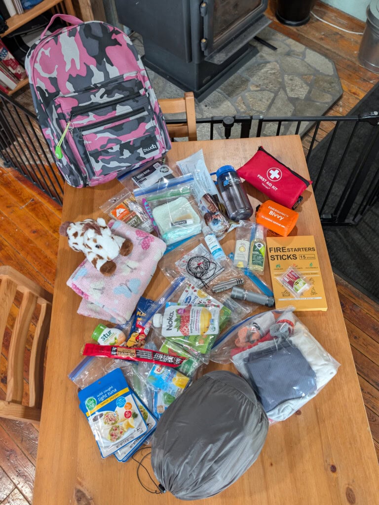 Flat lay of a kid-friendly 72-hour kit with a pink camo backpack, comfort blanket, stuffed animal, and emergency supplies on a wooden table.
