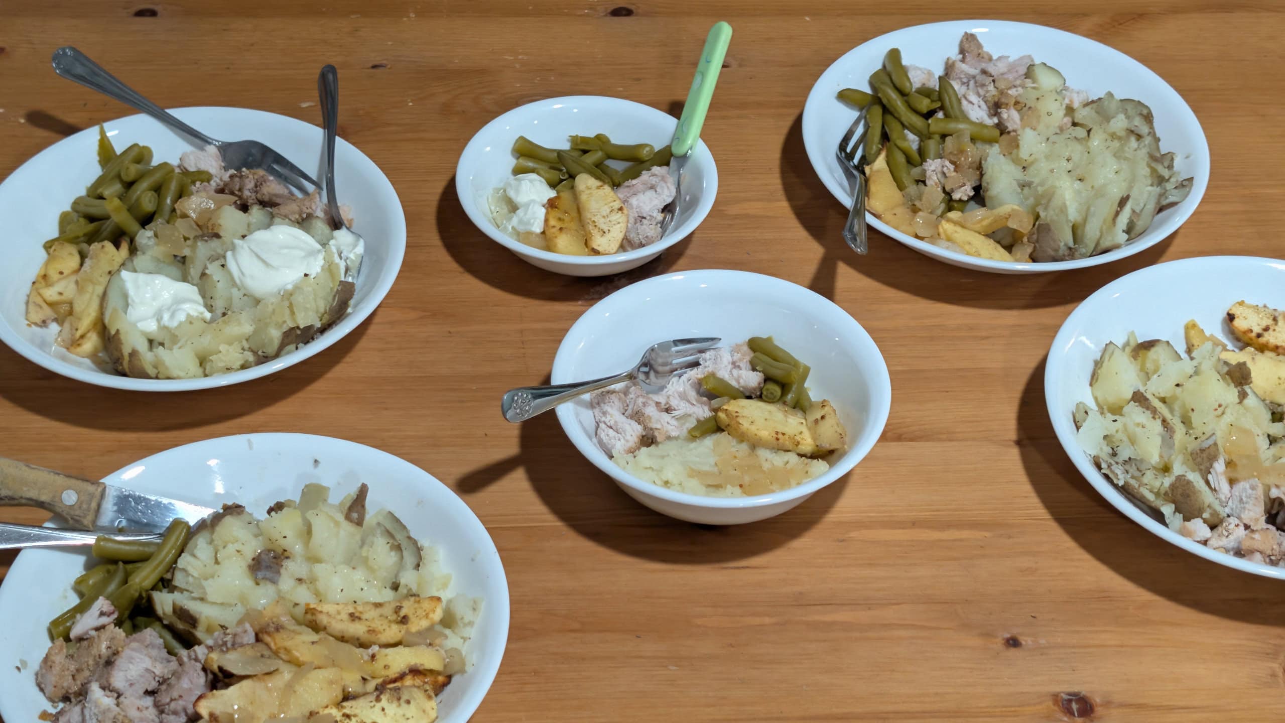 Six bowls of a frugal homestead meal with potatoes, green beans, and portioned meat on a rustic wooden table.