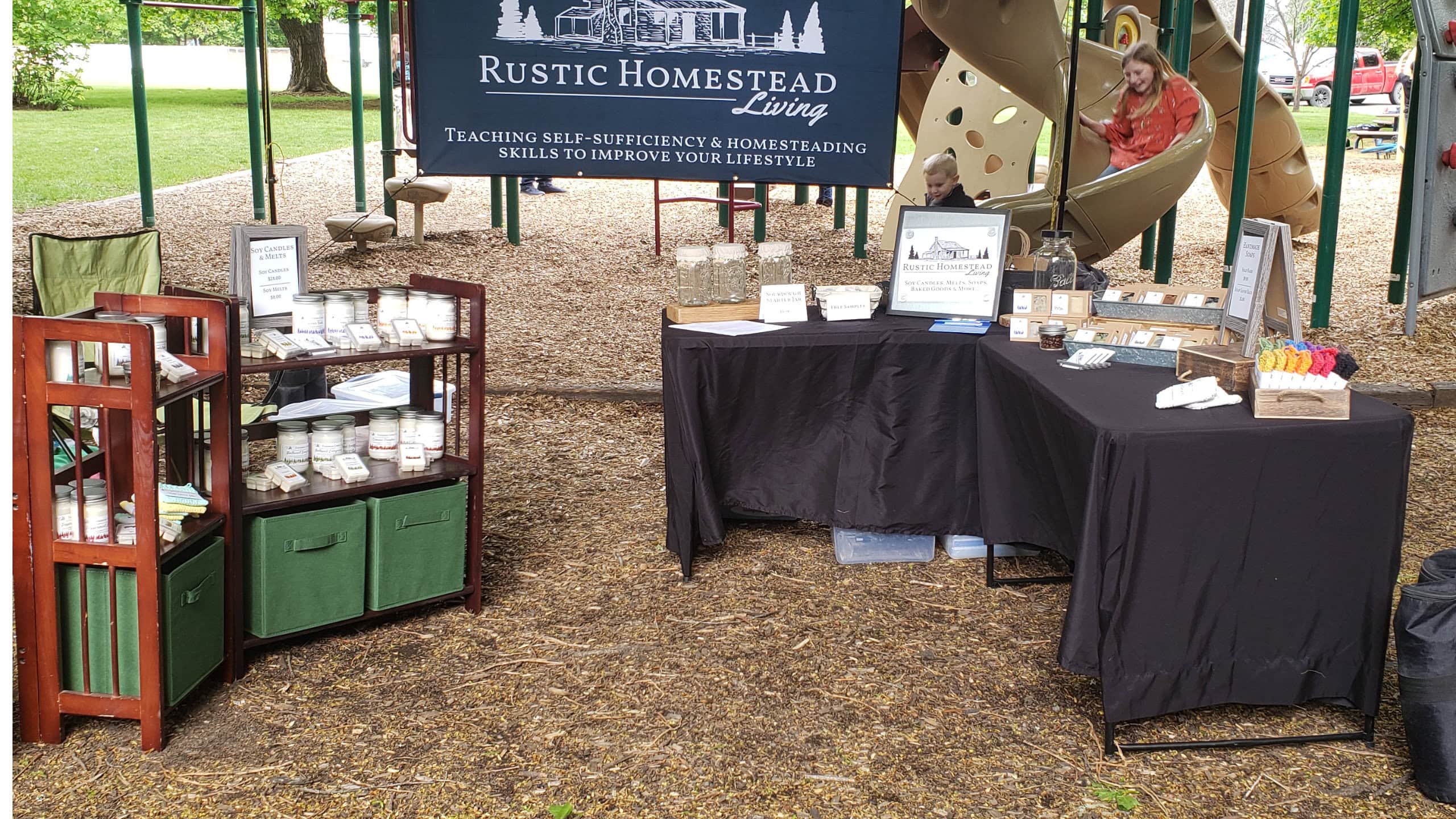 Farmers market booth under tent outside, products on tables and shelves.
