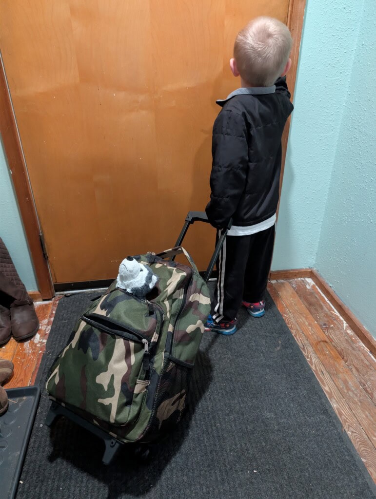 A three-year-old child standing at the front door holding a rolling 72-hour kit with a stuffed animal for comfort, practicing a family evacuation drill.